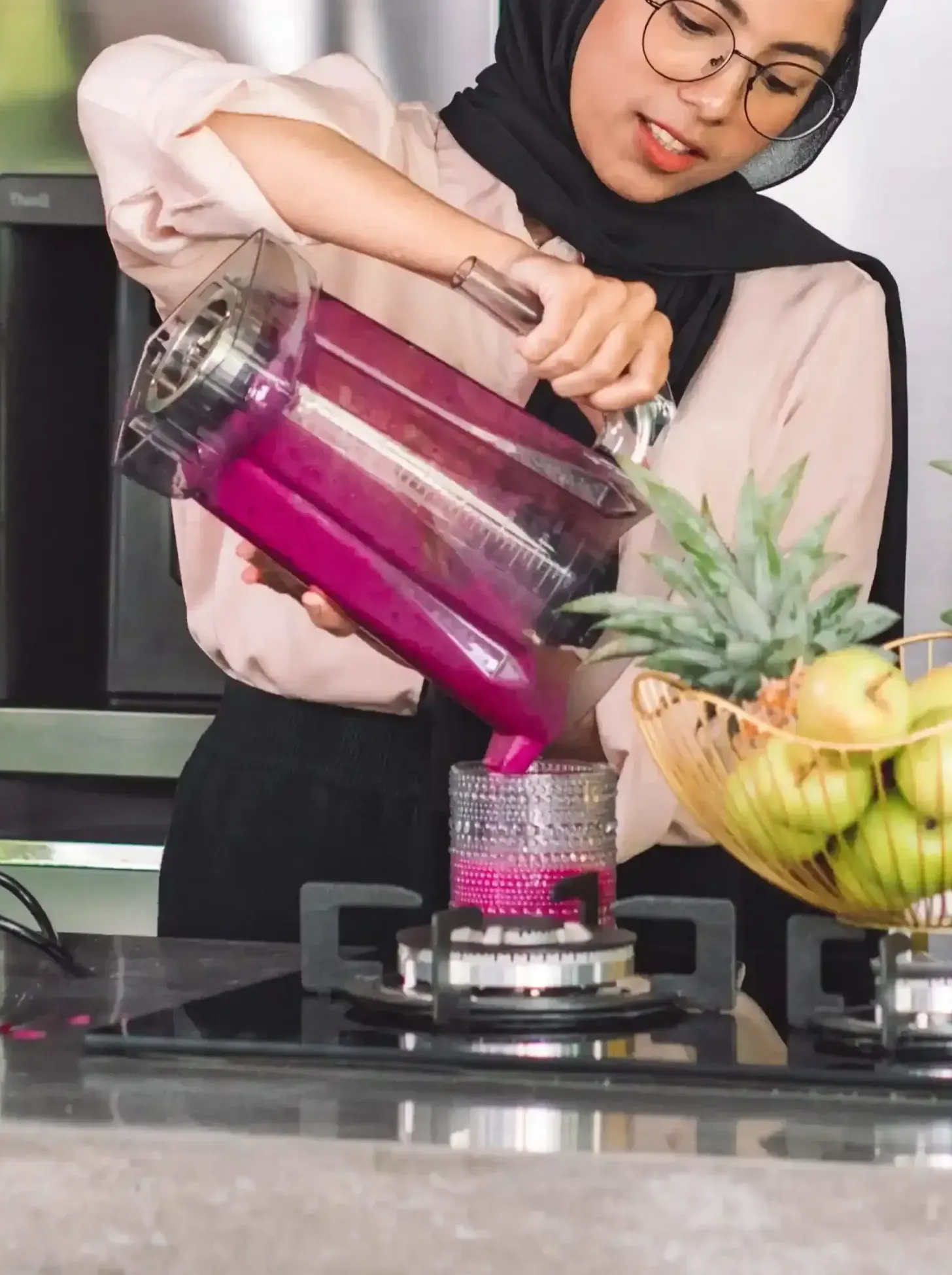 Wanita menuangkan cairan merah muda cerah dari blender ke dalam gelas; dapur dengan buah.