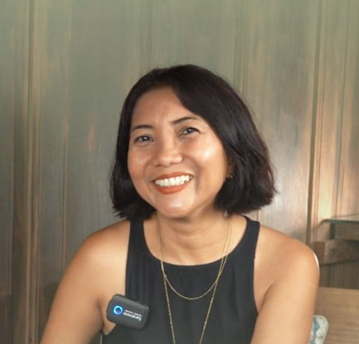 Seorang wanita mengenakan tank top hitam tersenyum sambil memegang mikrofon, dengan latar belakang kayu.