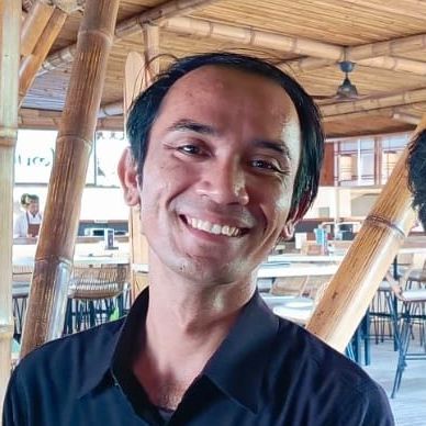 Orang yang tersenyum mengenakan kemeja hitam di sebuah kafe dengan deretan bambu.