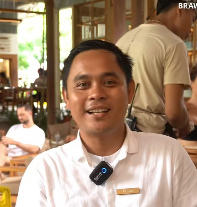 Seorang pria berbaju putih tersenyum sambil berbicara. Ia mengenakan mikrofon lavalier. Interior restoran.