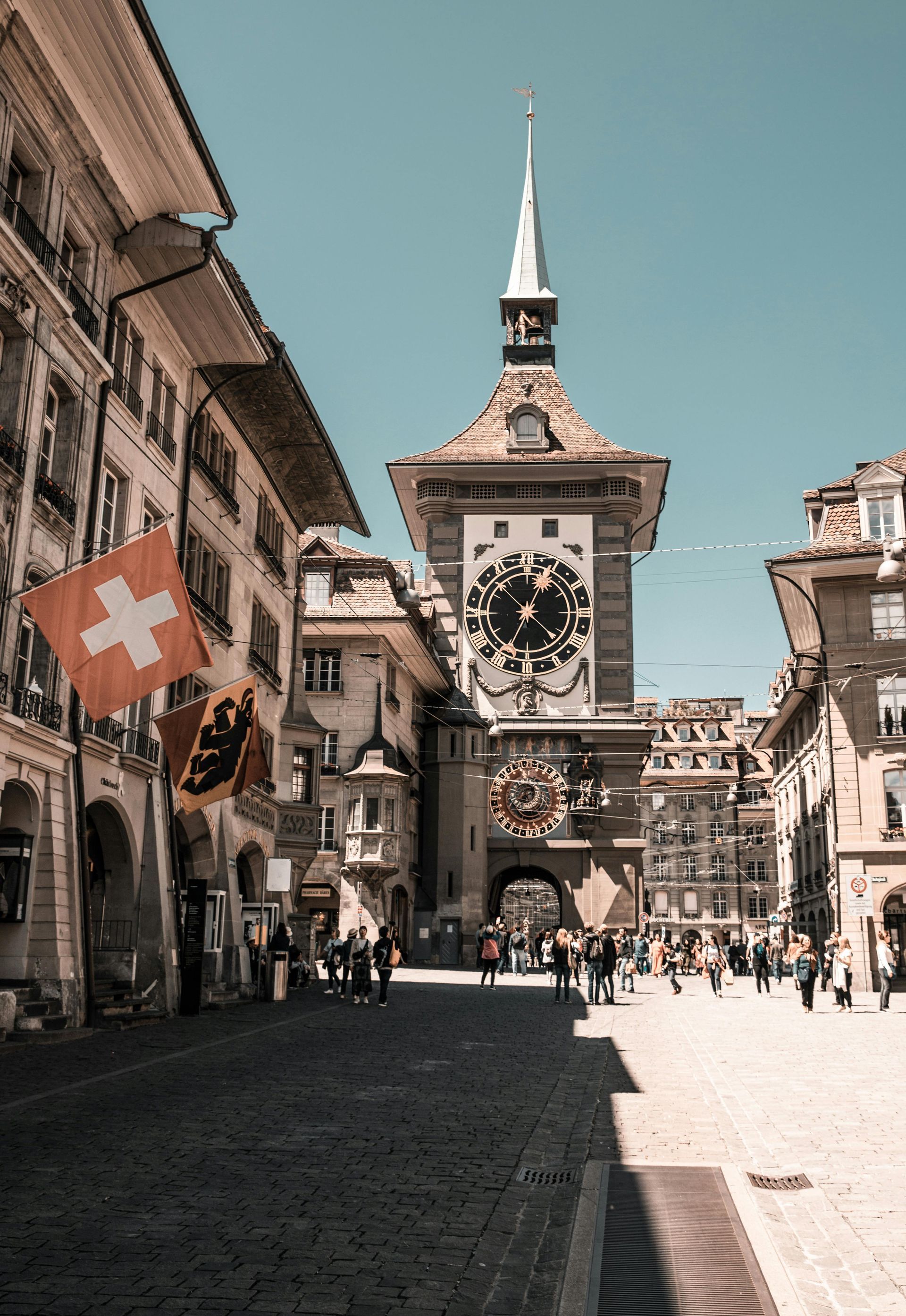 Menara jam di Bern, Swiss dengan bendera Swiss; orang-orang berjalan di jalan berbatu.