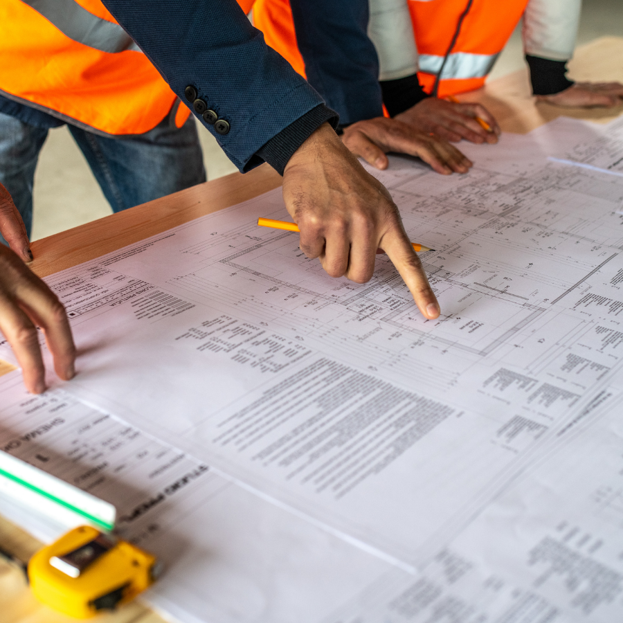 Construction workers in orange vests review blueprints on a table with a pencil and toy truck nearby