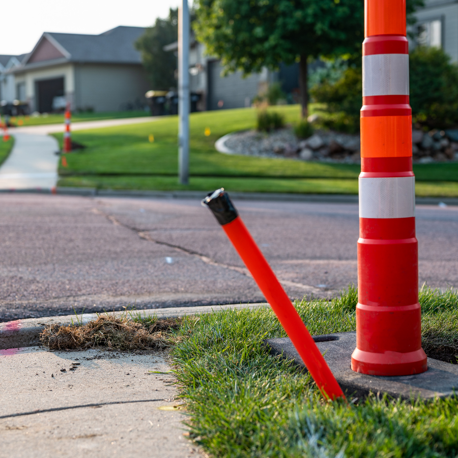 Red and white roadside delineator post leaning on a sidewalk beside a curb and grassy verge.