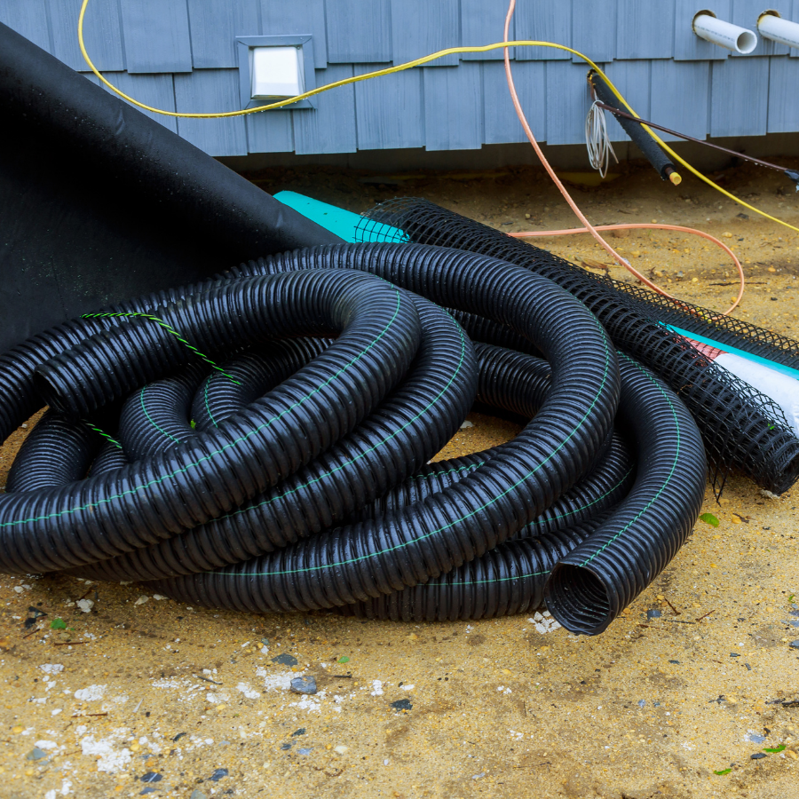 Coiled black corrugated drainage pipes on dirt beside a gray house wall