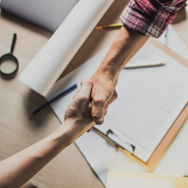 Two people shaking hands over a desk with papers, pens, and a notebook.