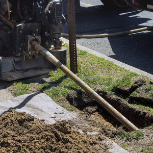 Construction drill digging a trench beside a sidewalk, with soil piled on the ground.