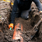 Worker digging up an orange pipe in muddy soil with a shovel and gloved hands