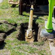 Yellow excavator arm digging a trench in a grassy yard beside a person in a green boot