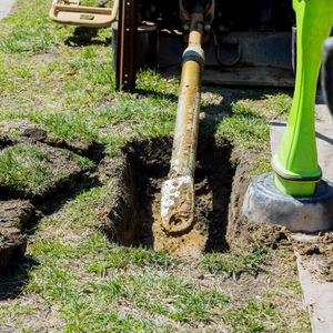 Yellow excavator arm digging a trench in a grassy yard beside a person in a green boot