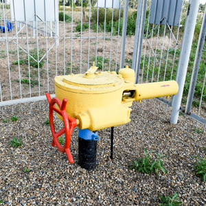 Yellow gas valve with red handwheel on gravel beside a metal fence