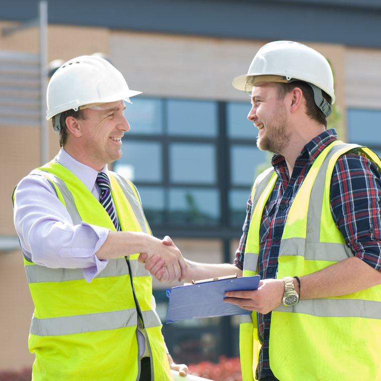 Two construction workers in hard hats and safety vests shake hands at a job site while holding plans.