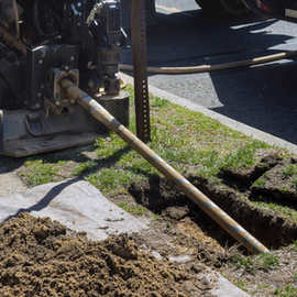 Utility drill rod boring into grass beside a sidewalk, with dirt piled up nearby.