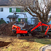 Red mini excavator digging a dirt trench beside a house with green shutters in a yard