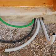 Several colored cables and a white pipe run through a concrete foundation into a gravel area.