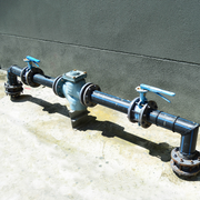 Black PVC pipe with blue-handled valves and a central gauge on a concrete floor against a gray wall