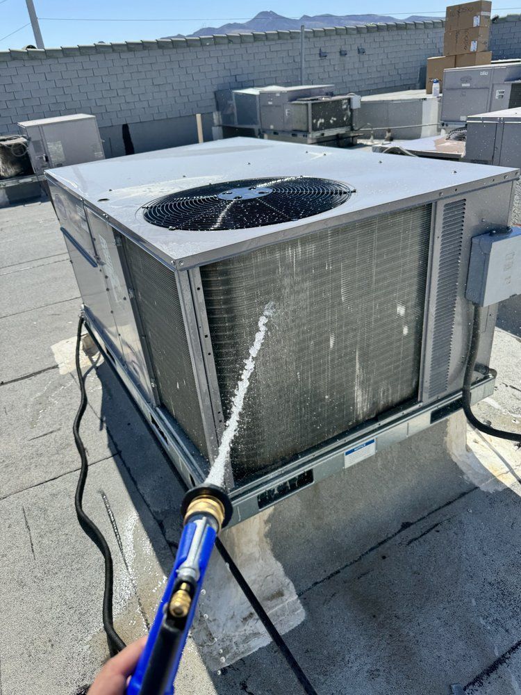 A hand sprays water from a hose onto the dirty metal coils of a rooftop HVAC unit.