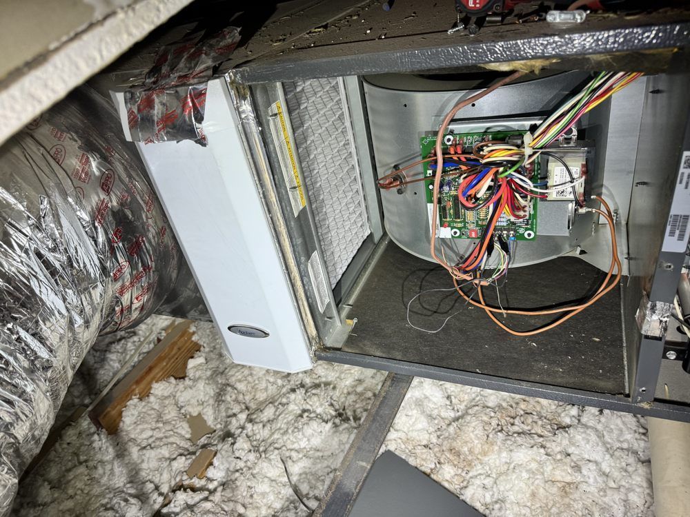An open HVAC blower compartment in an attic, showing a control board, wiring, and a white air filter.
