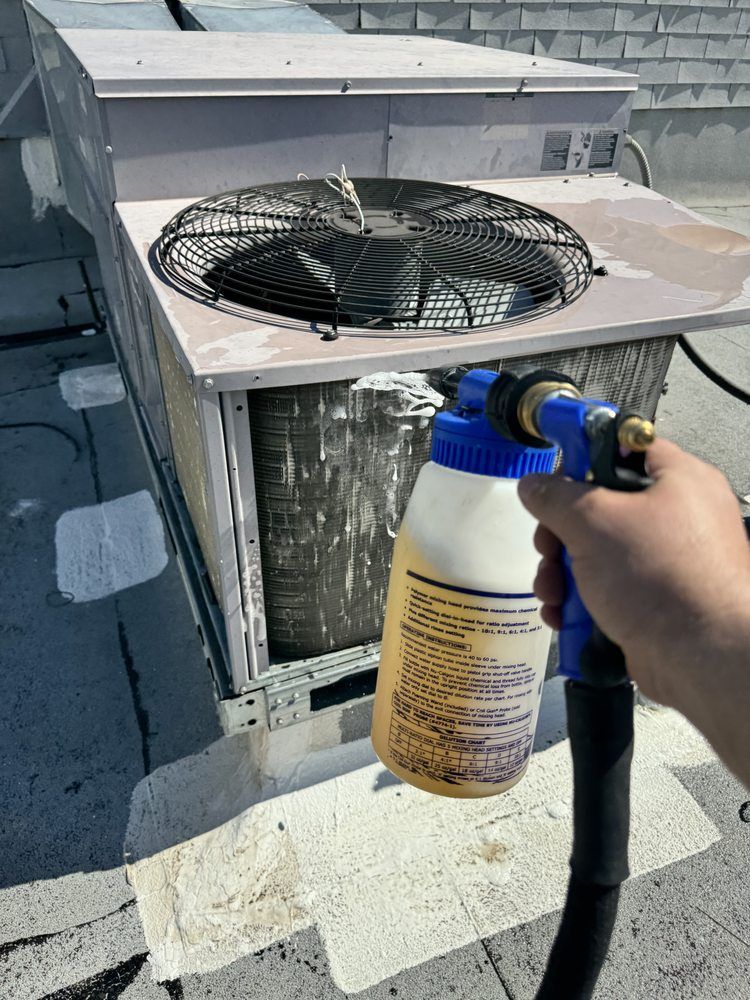 A hand uses a sprayer to apply cleaning foam to the metal cooling coils of an outdoor HVAC unit on a roof.