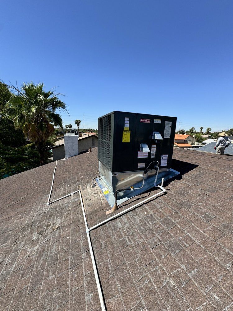 An HVAC rooftop unit sits on a shingled roof under a clear blue sky, with white piping running along the surface.