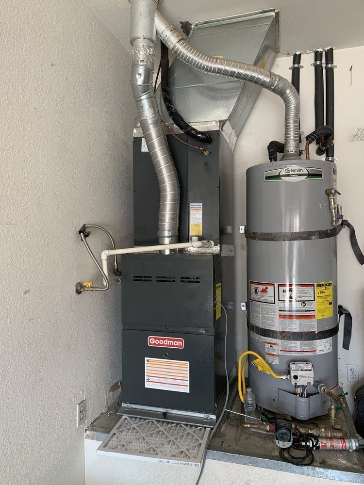 A gray HVAC furnace unit sits beside a large gray water heater in a corner, with silver metal ductwork connecting both.