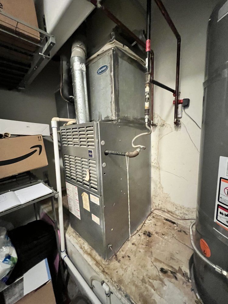 A gray HVAC furnace unit sits in a utility closet with nearby pipes, ductwork, and a water heater.