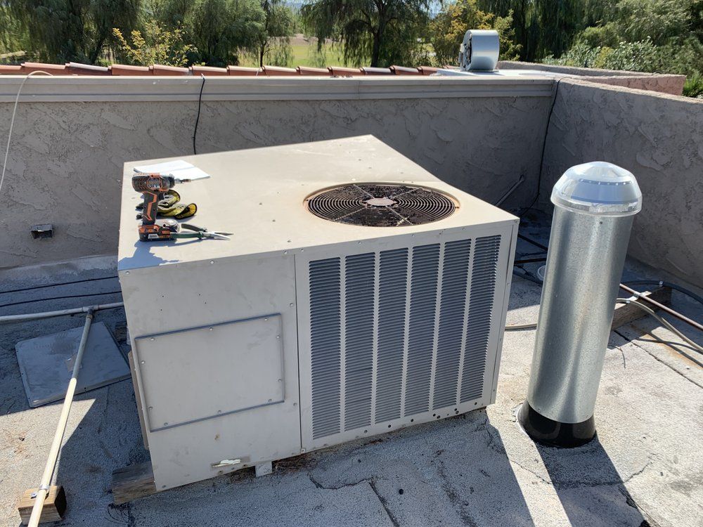 A roof-mounted air conditioning unit sitting next to a metal vent pipe, with a power drill resting on top.