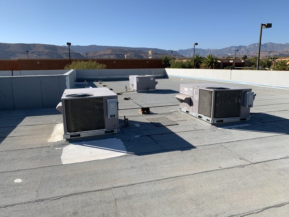 Three HVAC units sit on a flat, gray commercial rooftop under a clear blue sky, with mountains in the background.