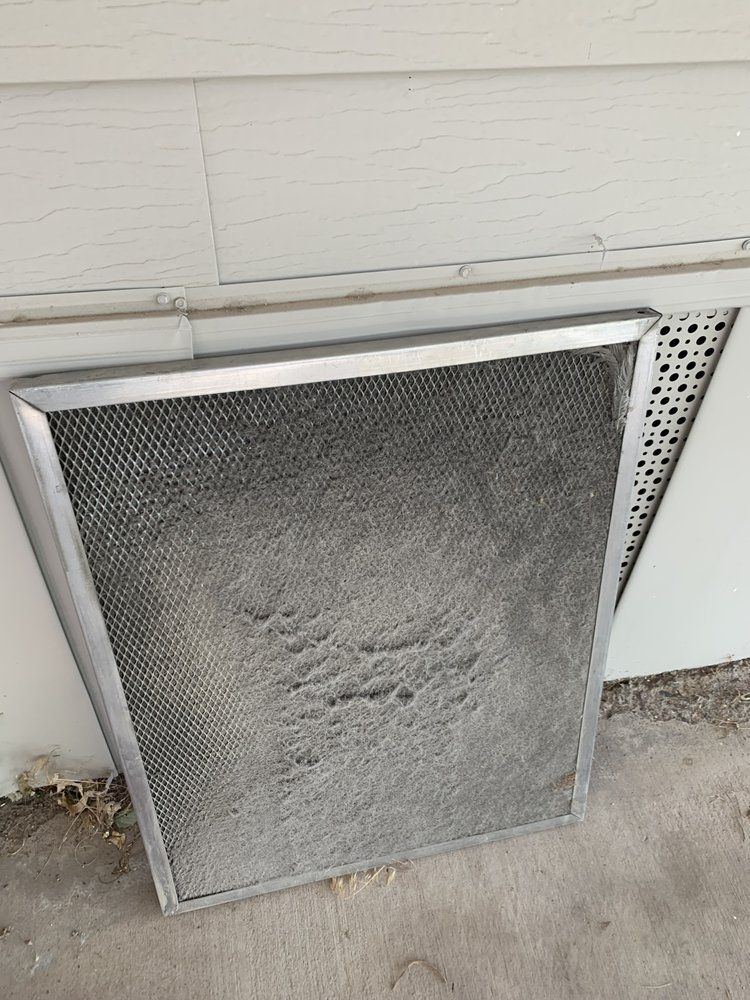 A dirty metal range hood grease filter leans against a white siding wall on a concrete surface.