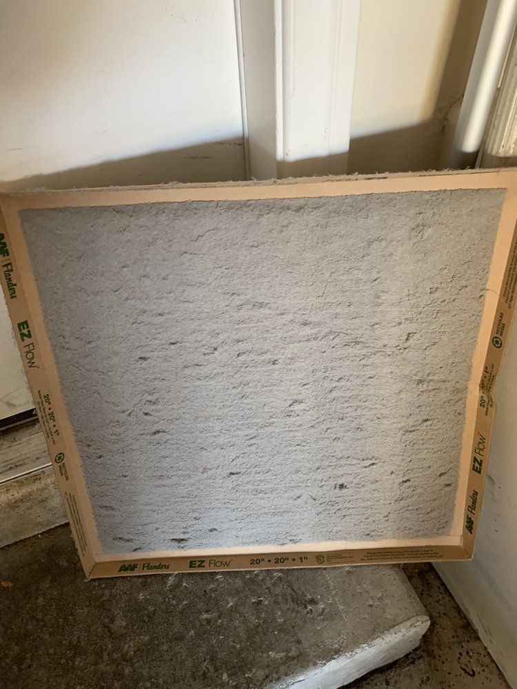 A dirty HVAC air filter with a cardboard frame leaning against a wall indoors.