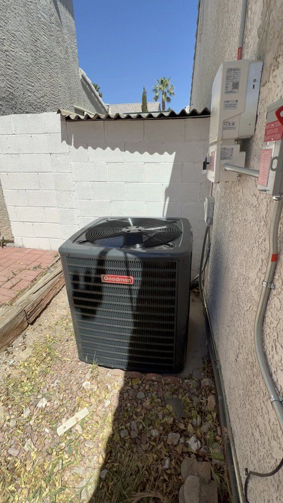 A gray HVAC unit sits on a gravel patch beside a beige stucco wall and a white block wall under a clear blue sky.