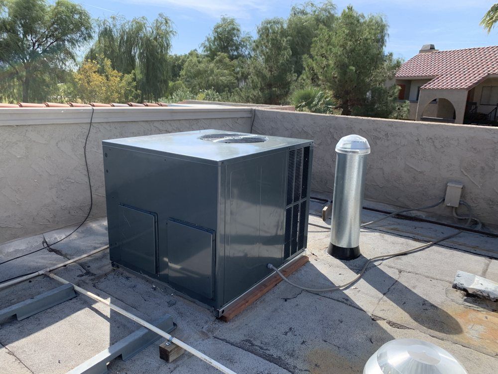 A grey HVAC rooftop unit sits on a flat, weathered roof next to a metal vent pipe, with trees and a house in the background.