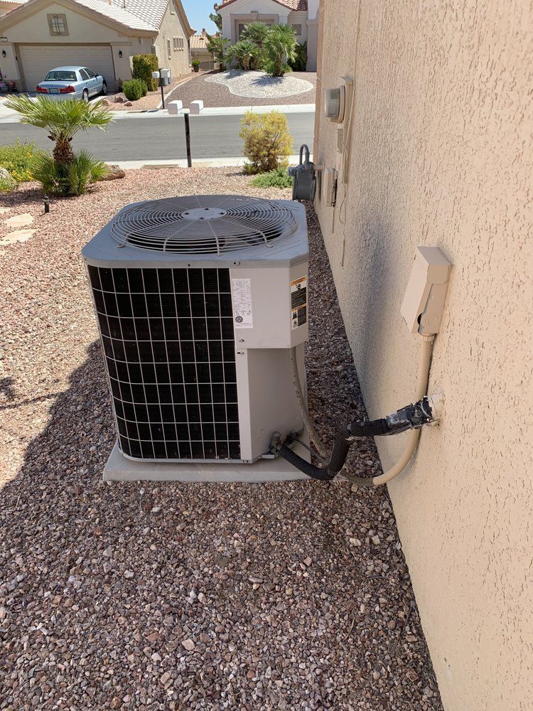A grey HVAC unit sits on a gravel patch beside a residential wall, connected by a power conduit and refrigerant lines.