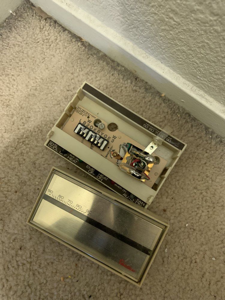 A detached beige mechanical thermostat with a metal faceplate lying on a carpeted floor.