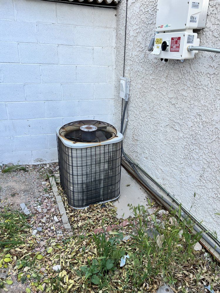 An outdoor HVAC condenser unit sits on a concrete pad next to a white stucco wall with an electrical disconnect box.