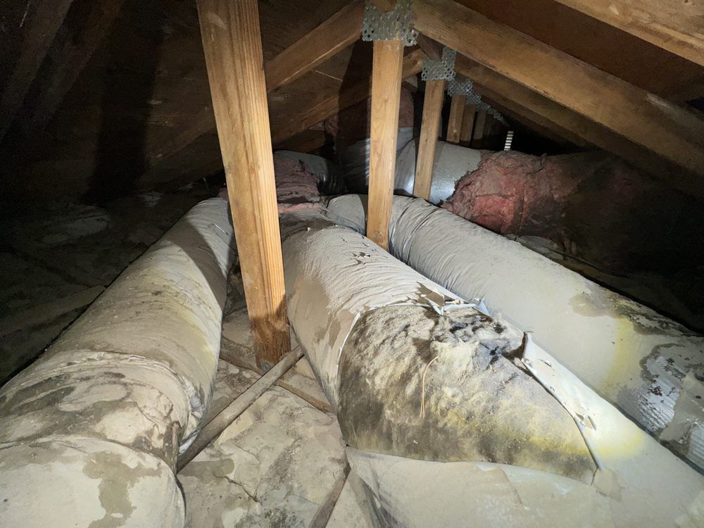 Damaged HVAC air ducts in a dark attic with wooden roof trusses and pink insulation visible in the background.