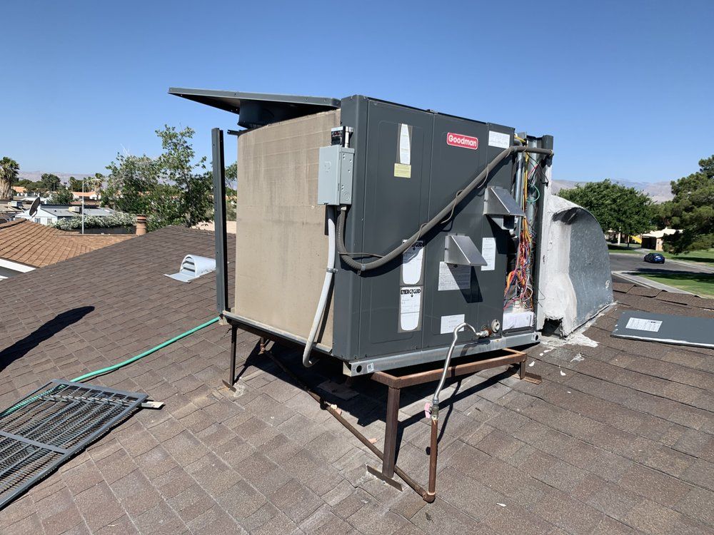 A dark-colored HVAC rooftop unit mounted on a metal stand on a shingled residential roof under a clear blue sky.