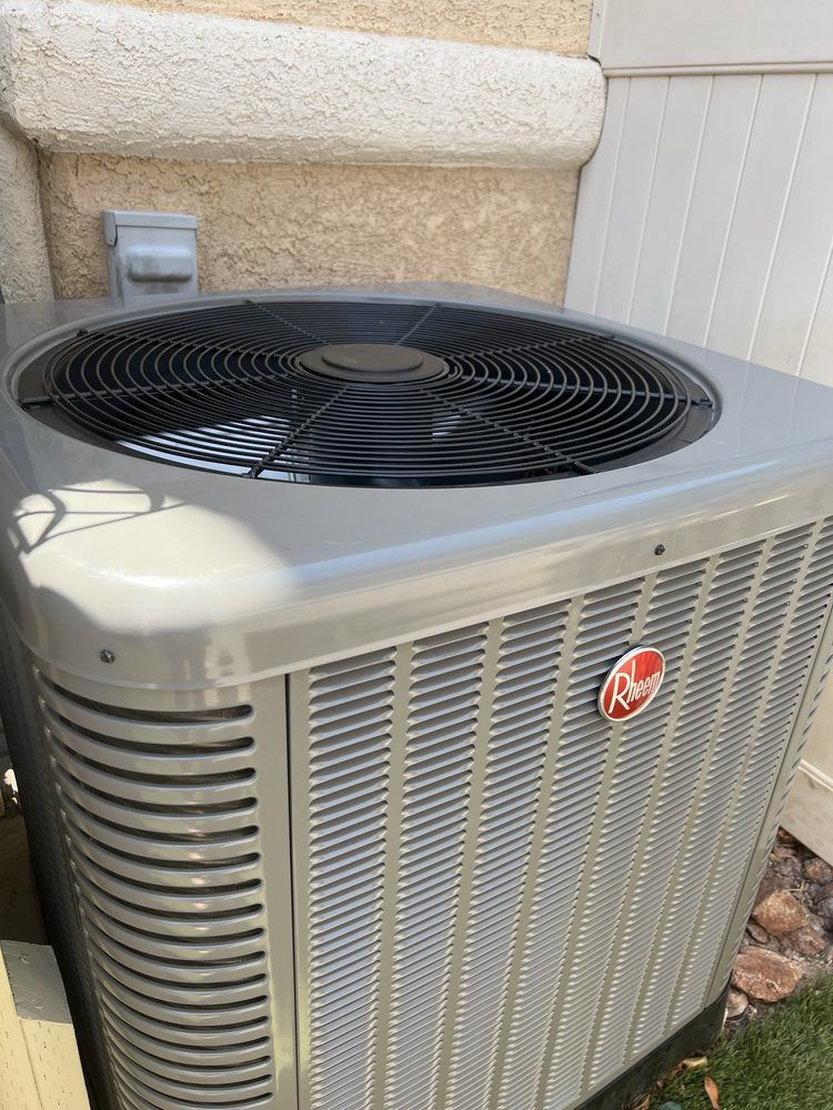 A silver Rheem air conditioning unit sits outdoors next to a light-colored stucco wall and a white fence.
