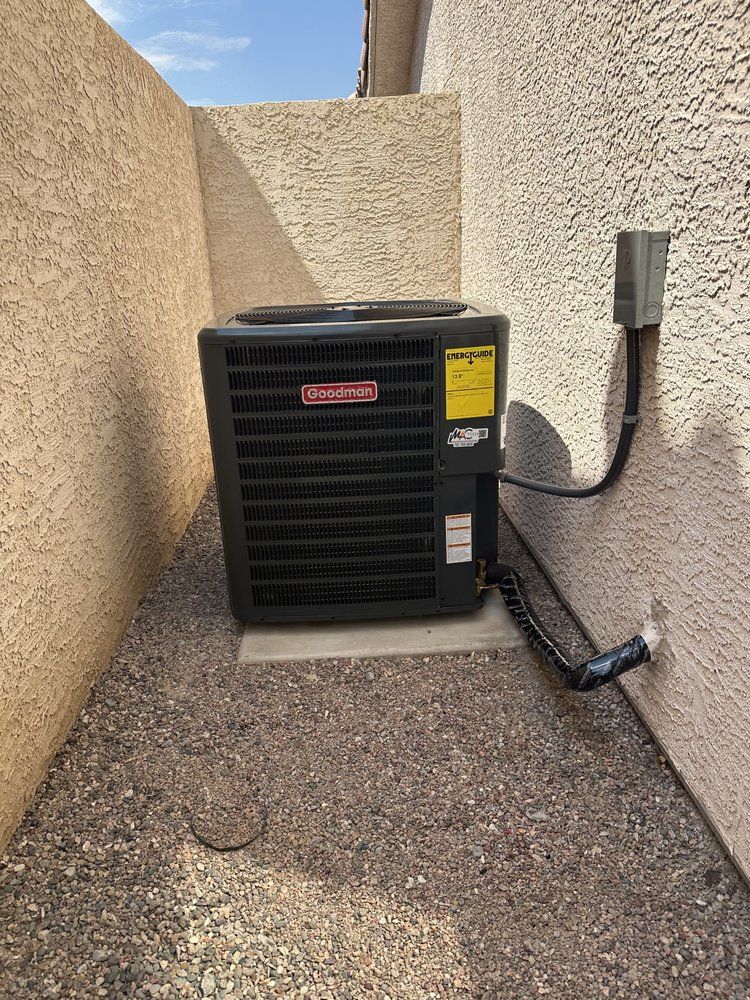 A dark-colored Goodman HVAC condenser unit sits on a gravel surface between two light-colored stucco walls.