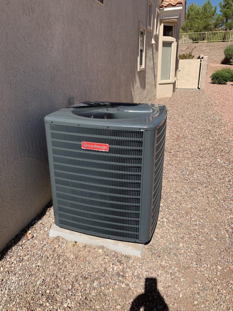 A grey Goodman brand air conditioning unit sits on a concrete pad next to the exterior wall of a stucco house.