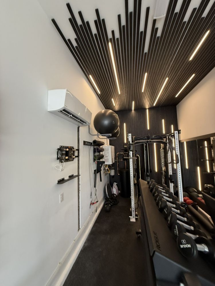 A modern home gym featuring black acoustic slat ceiling panels with recessed lighting, dumbbell racks, and an AC unit.