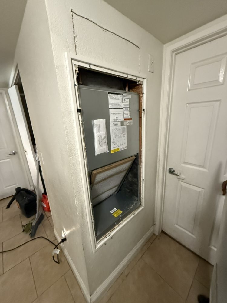 An HVAC air handler unit exposed behind an open wall panel in a hallway.