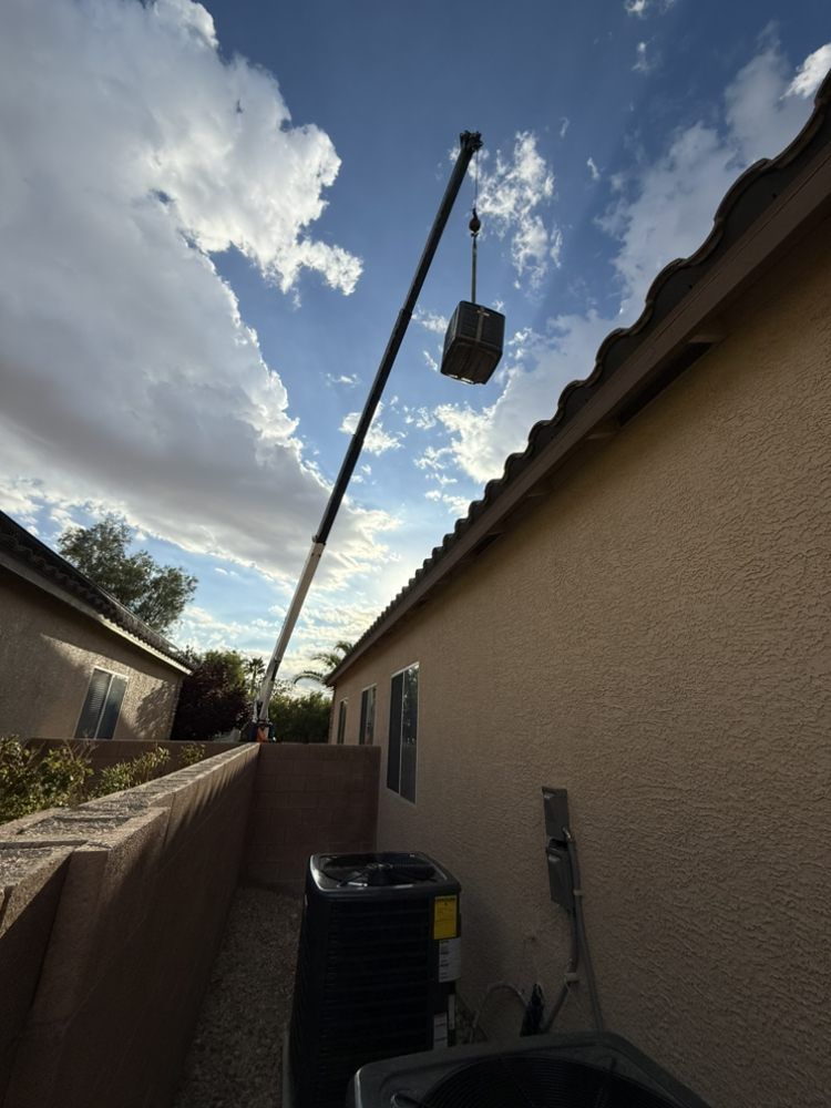 A crane lifts an HVAC unit above the roofline of a tan stucco home near a block wall and existing air conditioning units.