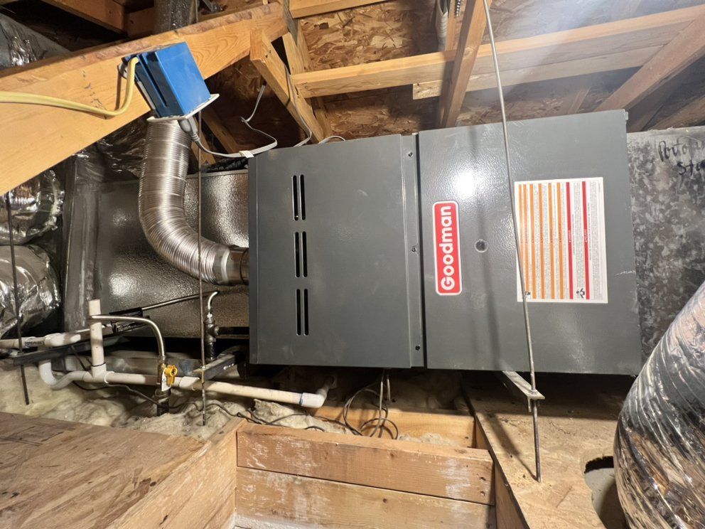 A gray Goodman HVAC furnace unit suspended by metal rods within an attic space with wooden rafters and ductwork.