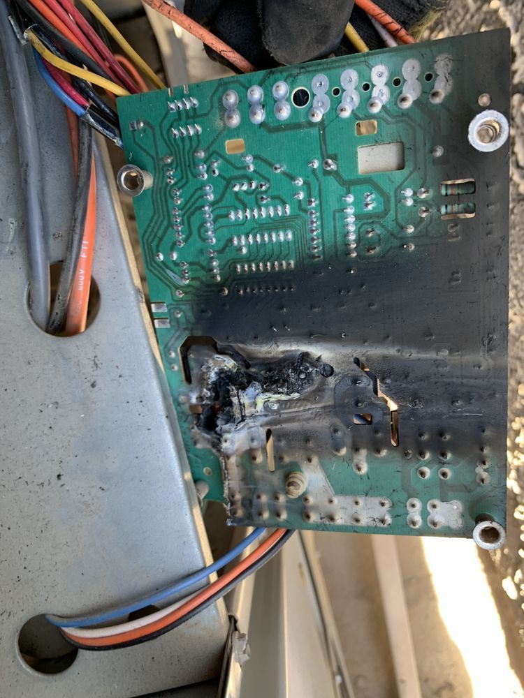 A green electronic circuit board with significant burn damage and charring on its lower half, surrounded by wires.
