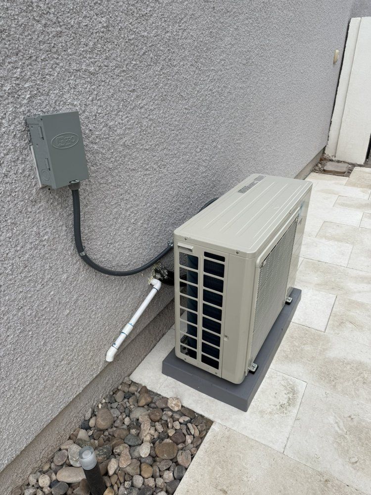 A beige outdoor mini-split heat pump unit sitting on a grey base next to a house wall with electrical and piping connections.