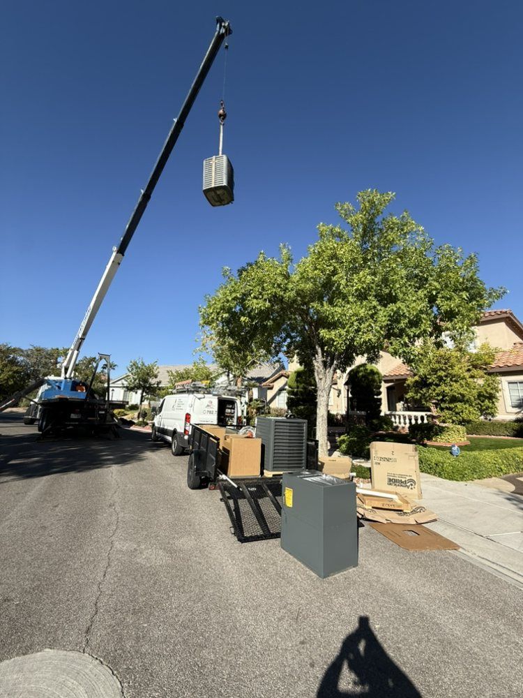 A crane lifts an HVAC unit into the air on a residential street next to a van and equipment on a trailer.