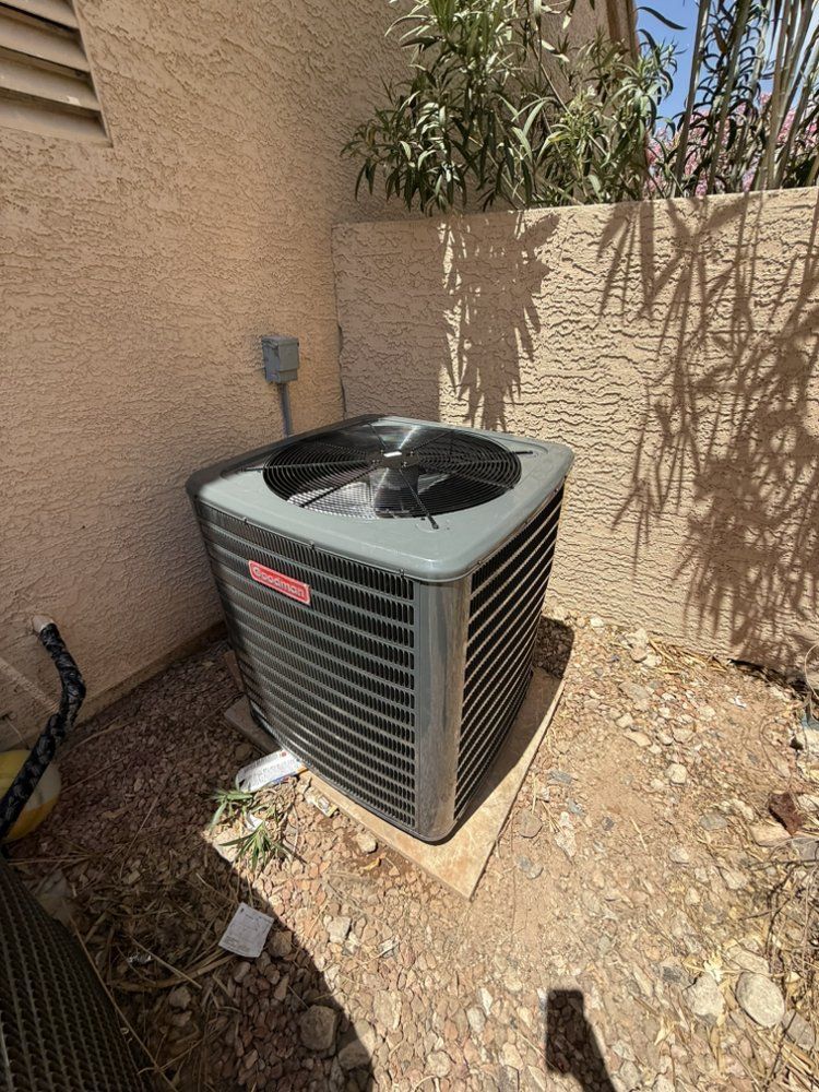 A grey Goodman air conditioning unit sits on a concrete slab in a rocky yard next to a stucco house wall.