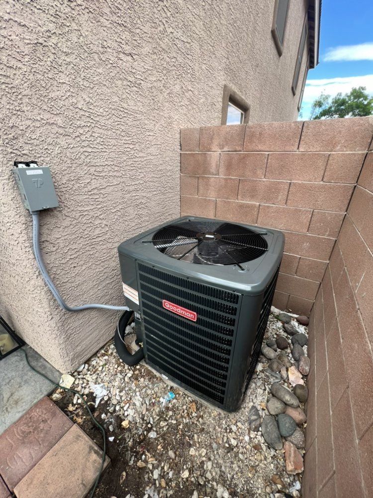A grey HVAC outdoor condenser unit sits on a gravel patch against a tan stucco wall next to a cinder block fence.