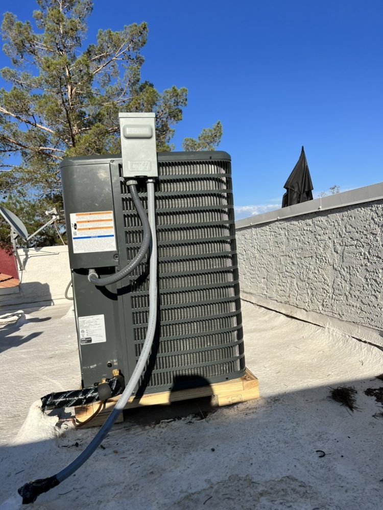 A gray air conditioning unit sitting on a flat roof under a clear blue sky, next to a textured white stucco wall.