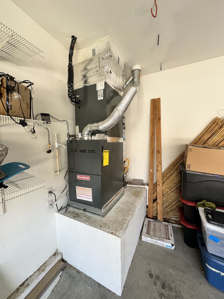 A Goodman HVAC furnace unit mounted on a white raised platform in a garage, with ductwork attached to the ceiling.